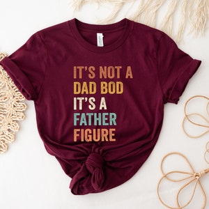 Pu&ograve; includere: Una maglietta color borgogna con il testo "It's not a dad bod it's a father figure" in un font in stile vintage.