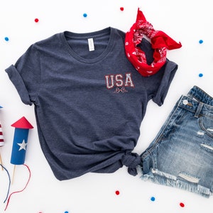 Puede incluir: Una camiseta azul marino con el texto "USA" en rojo, blanco y azul en la parte delantera. La camiseta se combina con pantalones cortos de mezclilla y una bandana roja.