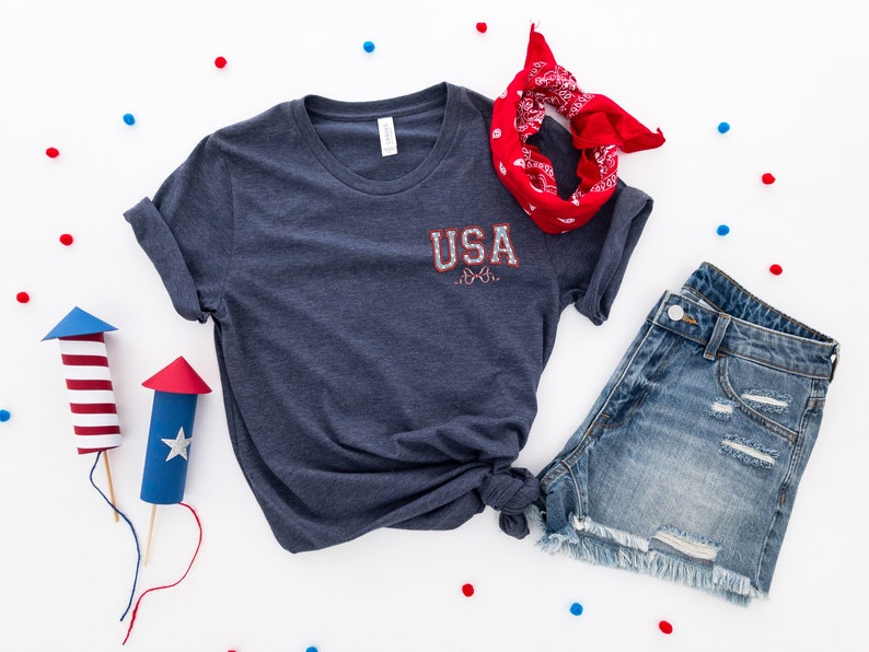 Puede incluir: Una camiseta azul marino con el texto "USA" en rojo, blanco y azul en la parte delantera. La camiseta se combina con pantalones cortos de mezclilla y una bandana roja.