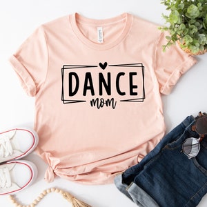 May include: A light pink t-shirt with the words "DANCE mom" in a black font, inside a black rectangular frame with a heart above the word "DANCE". The shirt is paired with blue denim shorts and white sneakers.