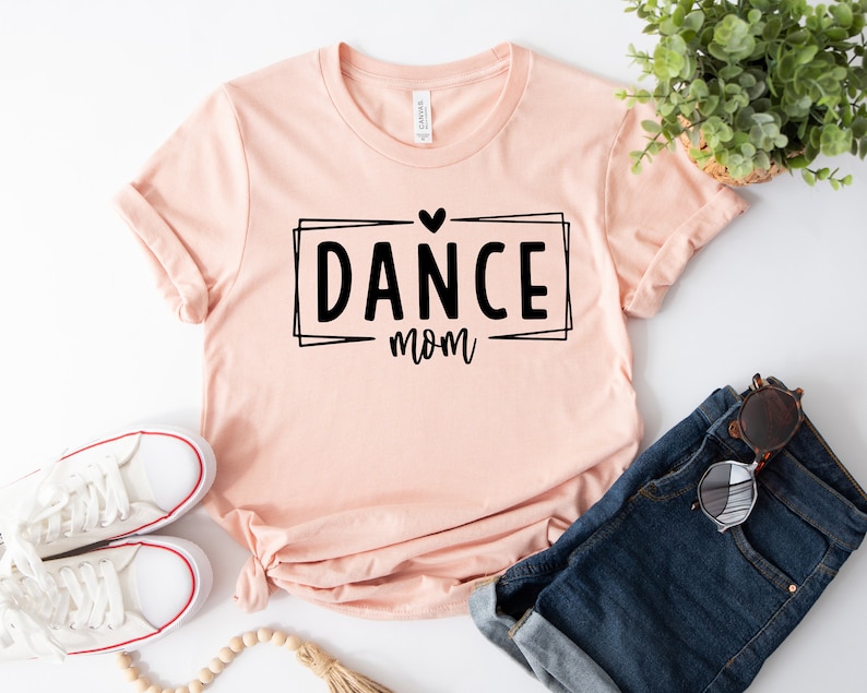 May include: A light pink t-shirt with the words "DANCE mom" in a black font, inside a black rectangular frame with a heart above the word "DANCE". The shirt is paired with blue denim shorts and white sneakers.