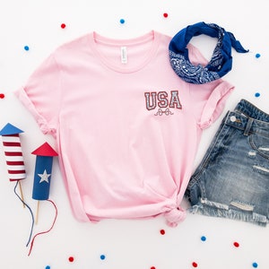 Puede incluir: Camiseta rosa con un gr&aacute;fico "USA" rojo, blanco y azul en el bolsillo delantero. La camiseta se combina con pantalones cortos de mezclilla y una bandana azul.