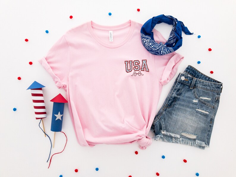 Puede incluir: Camiseta rosa con un gr&aacute;fico "USA" rojo, blanco y azul en el bolsillo delantero. La camiseta se combina con pantalones cortos de mezclilla y una bandana azul.