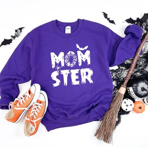K&ouml;nnte beinhalten: Lilafarbener Pullover mit dem Text "MOMSTER" in Wei&szlig;, mit einem Spinnennetz und einer Fledermaus. Der Pullover liegt auf einer wei&szlig;en Oberfl&auml;che mit einem Besen, Flederm&auml;usen und K&uuml;rbissen.