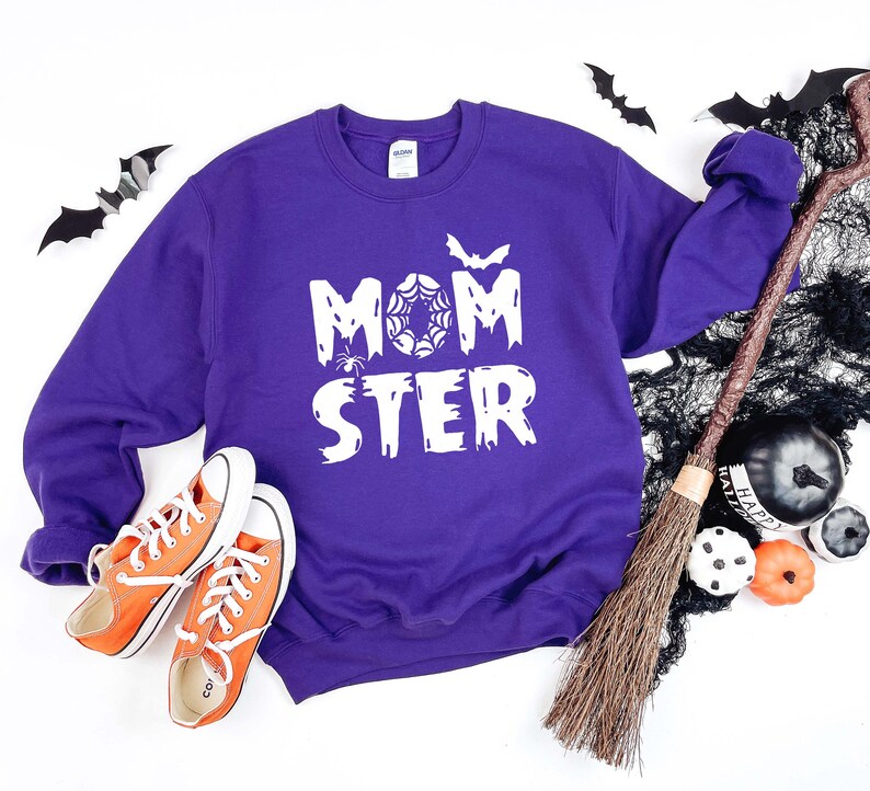 K&ouml;nnte beinhalten: Lilafarbener Pullover mit dem Text "MOMSTER" in Wei&szlig;, mit einem Spinnennetz und einer Fledermaus. Der Pullover liegt auf einer wei&szlig;en Oberfl&auml;che mit einem Besen, Flederm&auml;usen und K&uuml;rbissen.