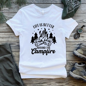 May include: White t-shirt with black text that reads "Life is better around the campfire". The text is surrounded by a campfire graphic with flames and logs, and pine trees.