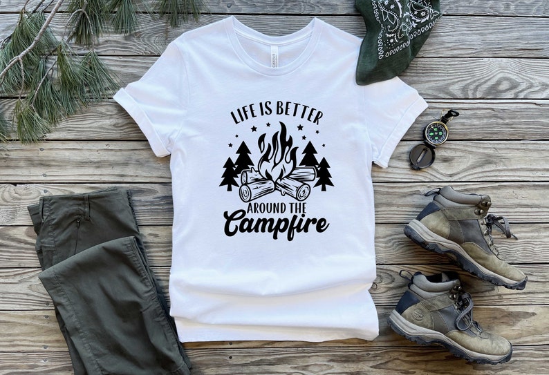 May include: White t-shirt with black text that reads "Life is better around the campfire". The text is surrounded by a campfire graphic with flames and logs, and pine trees.
