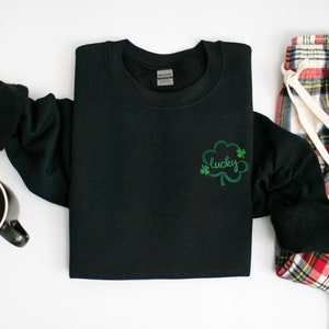 以下が含まれることがあります： 黒いスウェットシャツに、緑のクローバーと「lucky」という刺繍が施されています。