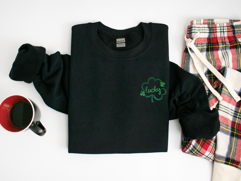 以下が含まれることがあります： 黒いスウェットシャツに、緑のクローバーと「lucky」という刺繍が施されています。