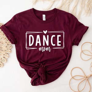 May include: A maroon t-shirt with the words "DANCE mom" in a white rectangular frame, with a small heart above the word "mom". The shirt is knotted at the bottom. The shirt is displayed on a white surface.
