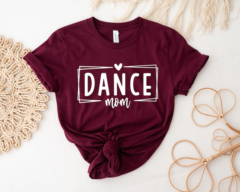 May include: A maroon t-shirt with the words "DANCE mom" in a white rectangular frame, with a small heart above the word "mom". The shirt is knotted at the bottom. The shirt is displayed on a white surface.