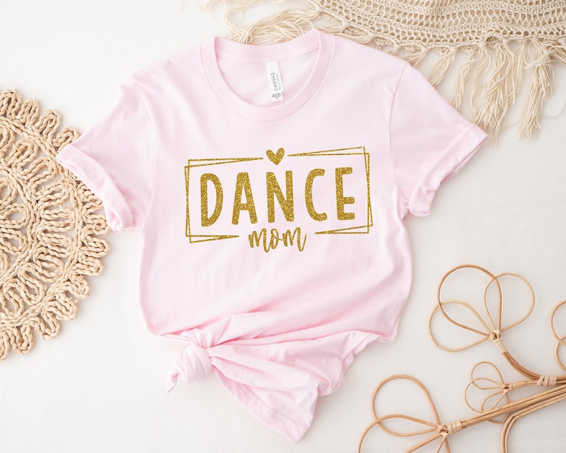 May include: A light pink t-shirt with the words "DANCE mom" in gold glitter lettering inside a gold rectangle. A small gold heart is above the word "DANCE". The shirt is knotted at the bottom and is displayed on a white surface.