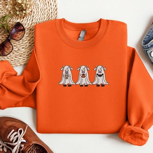 K&ouml;nnte beinhalten: Orangefarbener Pullover mit einem Motiv von drei wei&szlig;en Geisterk&uuml;hen, die Tassen Kaffee halten. Die K&uuml;he tragen Geisterkost&uuml;me mit wei&szlig;en Laken und haben schwarze Augen und H&ouml;rner.