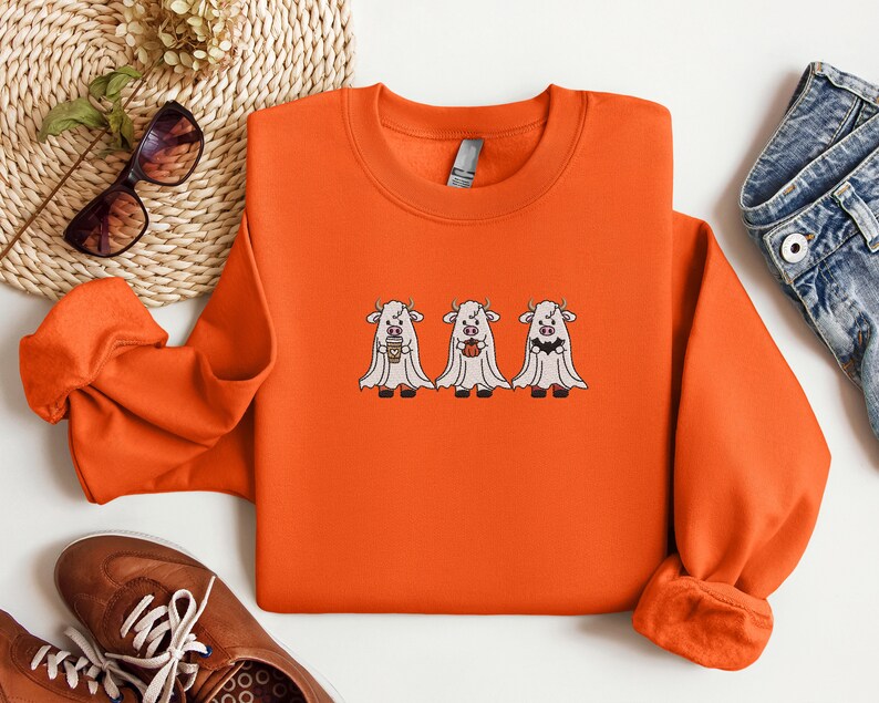 K&ouml;nnte beinhalten: Orangefarbener Pullover mit einem Motiv von drei wei&szlig;en Geisterk&uuml;hen, die Tassen Kaffee halten. Die K&uuml;he tragen Geisterkost&uuml;me mit wei&szlig;en Laken und haben schwarze Augen und H&ouml;rner.