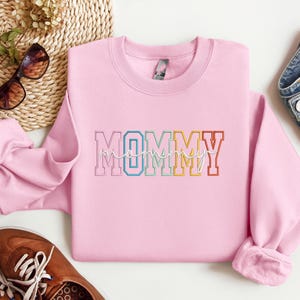 Pode incluir: Camisola cor-de-rosa com a palavra "MOMMY" em cores do arco-&iacute;ris com contorno branco.