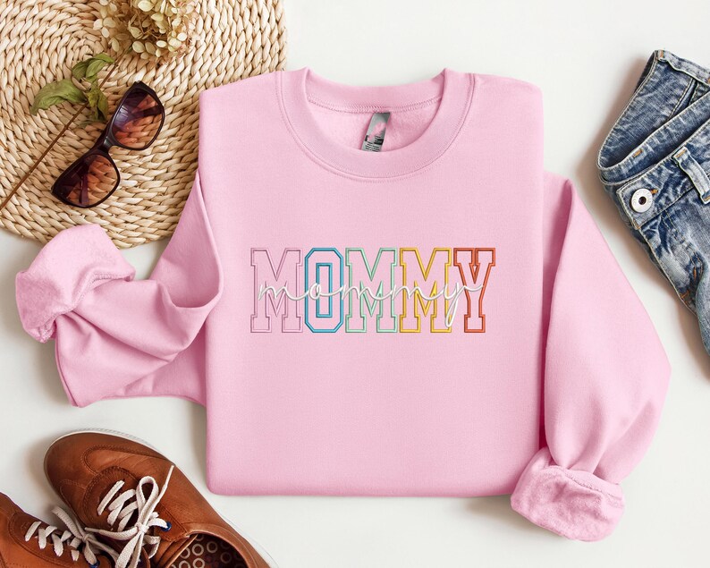 Pode incluir: Camisola cor-de-rosa com a palavra "MOMMY" em cores do arco-&iacute;ris com contorno branco.