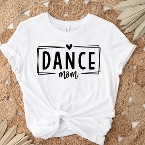 May include: White t-shirt with the text "DANCE mom" in a black, outlined rectangular box, topped with a heart. The shirt is knotted at the waist and rests on a woven, beige surface.
