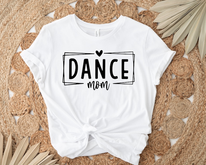 May include: White t-shirt with the text "DANCE mom" in a black, outlined rectangular box, topped with a heart. The shirt is knotted at the waist and rests on a woven, beige surface.
