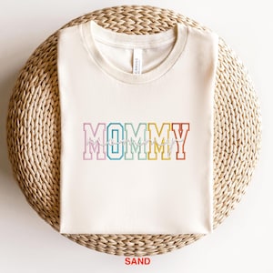 Pode incluir: Uma camiseta branca com a palavra "MOMMY" em cores do arco-&iacute;ris com uma fonte de escrita cursiva por baixo. A camiseta est&aacute; sobre uma superf&iacute;cie de palha tecida. A palavra "SAND" est&aacute; impressa em vermelho abaixo da camiseta.