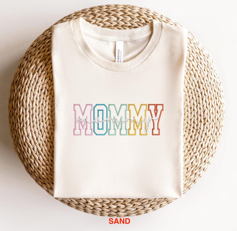Pode incluir: Uma camiseta branca com a palavra "MOMMY" em cores do arco-&iacute;ris com uma fonte de escrita cursiva por baixo. A camiseta est&aacute; sobre uma superf&iacute;cie de palha tecida. A palavra "SAND" est&aacute; impressa em vermelho abaixo da camiseta.