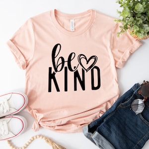 May include: A peach-colored t-shirt with the text "be kind" in black lettering with a small heart above the word "kind". The shirt is laid flat on a white surface with a pair of white sneakers, a pair of blue denim shorts, and a small potted plant.