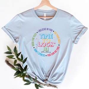 May include: Light blue t-shirt with the text "TIME TO LOCK IN" in a colorful design. The shirt also includes the phrases "I believe in you," "Do your best," "You can do this," and "Don't stress."