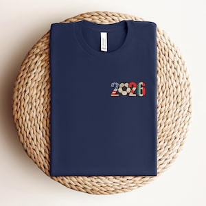 May include: Navy blue t-shirt with the number "2026" and a soccer ball graphic. The "2026" design incorporates the American flag, a soccer ball, and the colors red, green, and black. The shirt is displayed on a woven, circular surface.