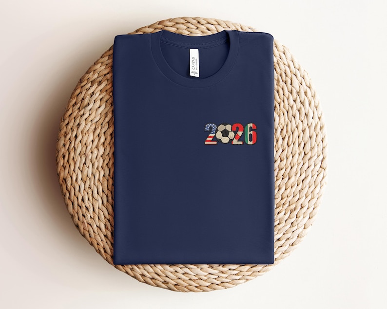 May include: Navy blue t-shirt with the number "2026" and a soccer ball graphic. The "2026" design incorporates the American flag, a soccer ball, and the colors red, green, and black. The shirt is displayed on a woven, circular surface.