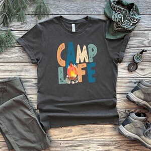 K&ouml;nnte beinhalten: Ein dunkelgraues T-Shirt mit dem Text "Camp Life" in bunten, Retro-Lettern. Ein Lagerfeuer mit Flammen befindet sich in der Mitte des Textes. Das Shirt liegt auf einer Holzoberfl&auml;che mit einem Bandana, einem Kompass und Wanderschuhen.