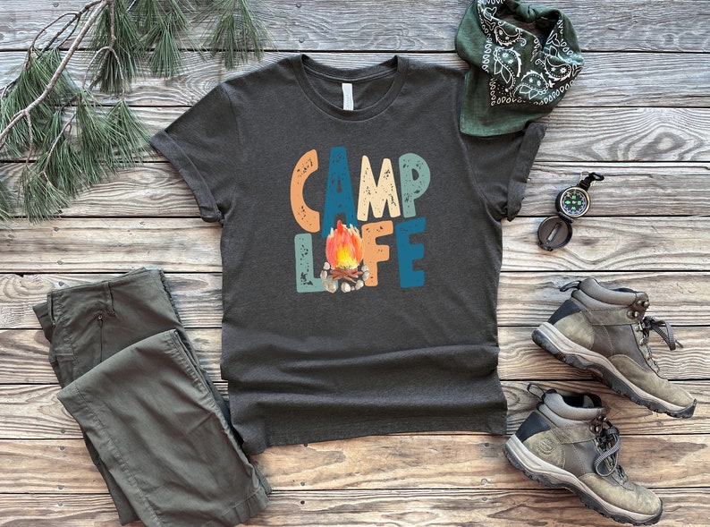 K&ouml;nnte beinhalten: Ein dunkelgraues T-Shirt mit dem Text "Camp Life" in bunten, Retro-Lettern. Ein Lagerfeuer mit Flammen befindet sich in der Mitte des Textes. Das Shirt liegt auf einer Holzoberfl&auml;che mit einem Bandana, einem Kompass und Wanderschuhen.