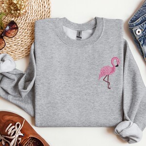 May include: A gray sweatshirt with a pink flamingo embroidered on the front. The sweatshirt is a crew neck style and has a relaxed fit.