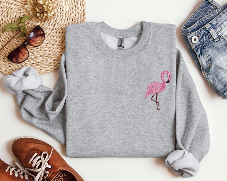 May include: A gray sweatshirt with a pink flamingo embroidered on the front. The sweatshirt is a crew neck style and has a relaxed fit.