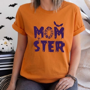 K&ouml;nnte beinhalten: Orangefarbenes T-Shirt mit dem Text "MOMSTER" in Lila mit einer Spinne, einem Spinnennetz und einer Fledermausgrafik.