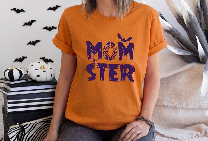 K&ouml;nnte beinhalten: Orangefarbenes T-Shirt mit dem Text "MOMSTER" in Lila mit einer Spinne, einem Spinnennetz und einer Fledermausgrafik.