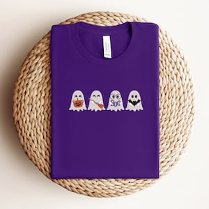May include: Purple t-shirt with a row of five embroidered ghosts. The ghosts have different expressions, including a pumpkin face, a smiling face, a ghost with glasses, a ghost with a bat, and a ghost with a frown.