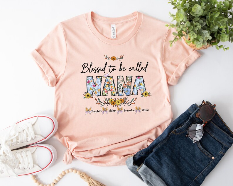 Op de afbeelding: Perzikkleurige T-shirt met de tekst "Blessed to be called NANA" in zwarte letters met bloemmotieven. De namen "Sophia, Alex, Brandon, Olive" staan in het zwart onder de tekst.