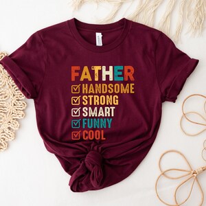 K&ouml;nnte beinhalten: Ein maroonfarbenes T-Shirt mit dem Text "FATHER" in einer Vintage-Schriftart. Unter dem Text befindet sich eine Checkliste mit folgenden Punkten: "HANDSOME", "STRONG", "SMART", "FUNNY" und "COOL". Jeder Punkt ist mit einem gr&uuml;nen H&auml;kchen abgehakt.