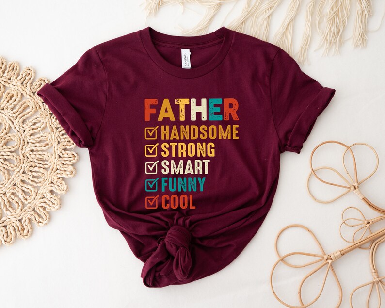 K&ouml;nnte beinhalten: Ein maroonfarbenes T-Shirt mit dem Text "FATHER" in einer Vintage-Schriftart. Unter dem Text befindet sich eine Checkliste mit folgenden Punkten: "HANDSOME", "STRONG", "SMART", "FUNNY" und "COOL". Jeder Punkt ist mit einem gr&uuml;nen H&auml;kchen abgehakt.