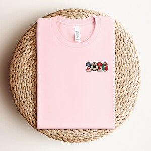 May include: A light pink t-shirt with a small graphic of the number "2026" and a soccer ball design. The graphic incorporates the colors of various national flags. The shirt is folded and placed on a woven, circular surface.
