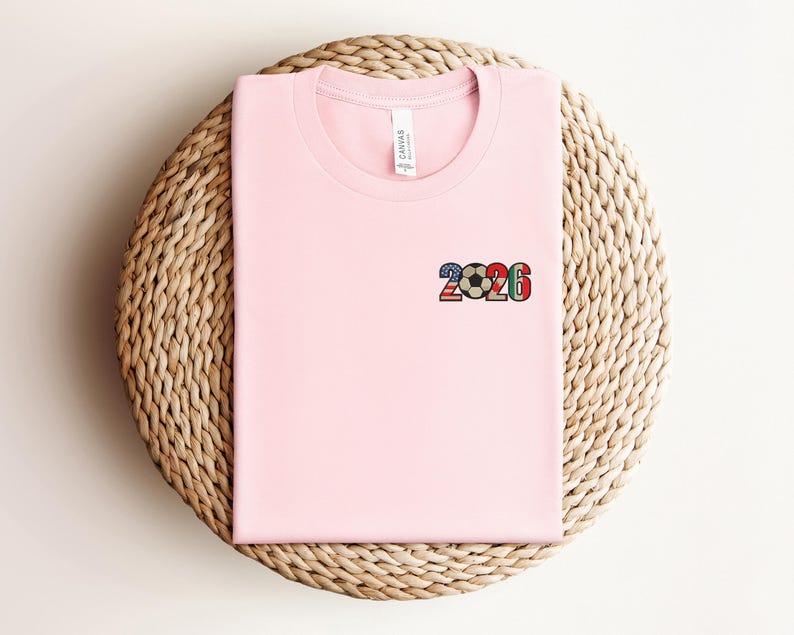 May include: A light pink t-shirt with a small graphic of the number "2026" and a soccer ball design. The graphic incorporates the colors of various national flags. The shirt is folded and placed on a woven, circular surface.
