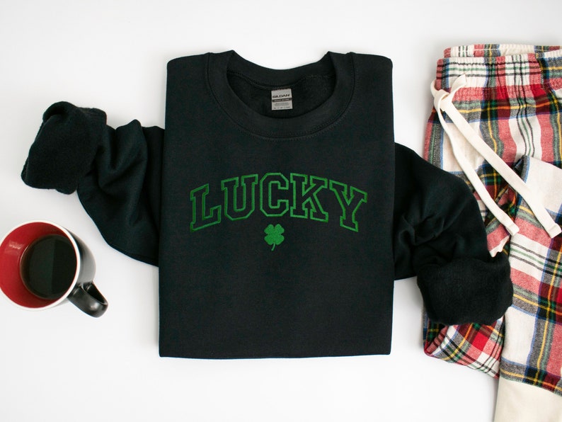 以下が含まれることがあります： 緑のクローバーと、その上に緑色のアーチ状に「LUCKY」と書かれた黒いスウェットシャツ