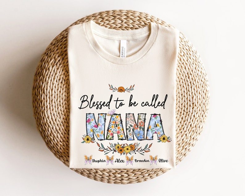 Op de afbeelding: Een wit T-shirt met de tekst "Blessed to be called NANA" in kleurrijke bloemenletters. De namen Sophia, Alex, Brandon en Olive staan onder de tekst met vlinderillustraties.