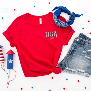 Puede incluir: Camiseta roja con un gr&aacute;fico blanco de "USA" en la parte delantera, pantalones cortos de mezclilla azul, un pa&ntilde;uelo azul y confeti rojo y azul sobre un fondo blanco.