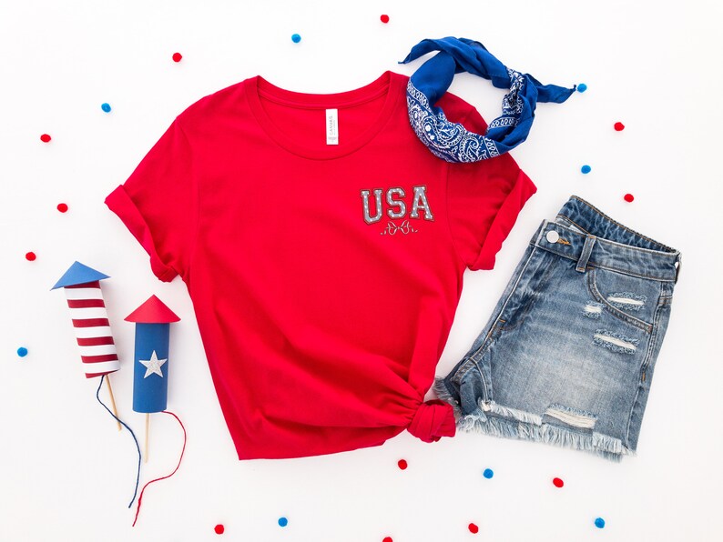 Puede incluir: Camiseta roja con un gr&aacute;fico blanco de "USA" en la parte delantera, pantalones cortos de mezclilla azul, un pa&ntilde;uelo azul y confeti rojo y azul sobre un fondo blanco.