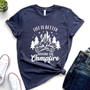 May include: A navy blue t-shirt with a white graphic design of a campfire with trees and stars. The text on the shirt reads "Life is better around the campfire".
