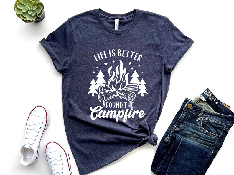 May include: A navy blue t-shirt with a white graphic design of a campfire with trees and stars. The text on the shirt reads "Life is better around the campfire".