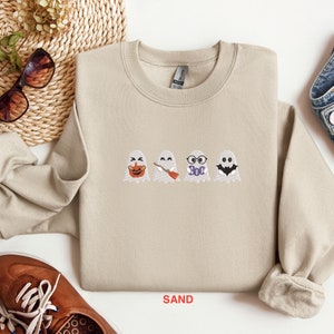 May include: A sand-colored sweatshirt with a crew neck and long sleeves. The sweatshirt features an embroidered design of four ghosts with different expressions and accessories. The ghosts are wearing a pumpkin, a broom, glasses that say "Boo", and a bat.