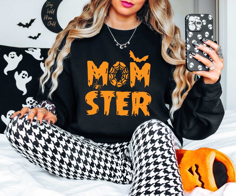 K&ouml;nnte beinhalten: Schwarzer Pullover mit orangefarbenem Text, der "Momster" mit einem Spinnennetz- und Fledermaus-Motiv lautet. Das Model tr&auml;gt schwarz-wei&szlig; karierte Leggings und orangefarbene Hausschuhe.