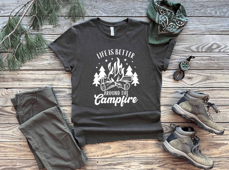 May include: A dark gray t-shirt with white text that reads "Life is better around the campfire". The text is surrounded by a campfire graphic with flames and trees.