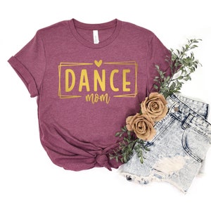 May include: A maroon t-shirt with the words "DANCE mom" in gold lettering inside a gold rectangle. The shirt is styled with denim shorts, faux flowers, and greenery on a white background.